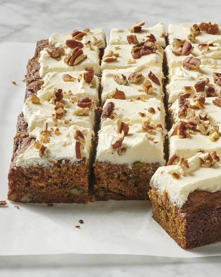 cut zucchini cake topped with cream cheese frosting and topped with chopped nuts on parchment paper on a marble surface