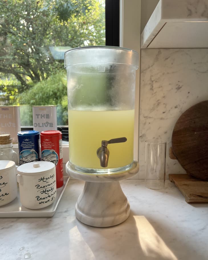 Glass beverage dispenser with yellow liquid on a marble stand, next to spice jars and canned drinks on a kitchen counter.