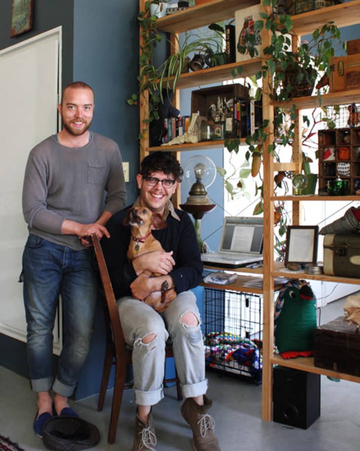 Two people in a home office with a dog, surrounded by plants, books, and a laptop on a wooden shelf.