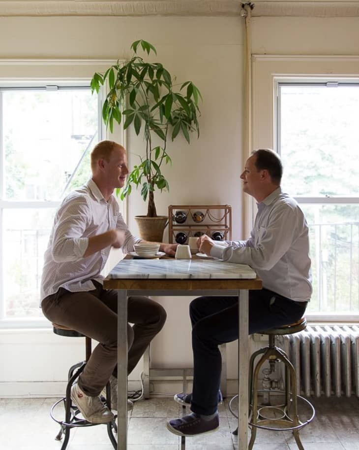 Two men sitting at a high table with coffee cups, near a large plant and window.