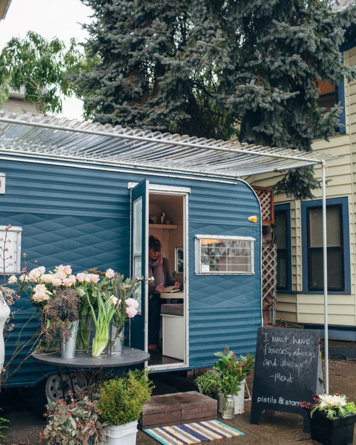 Blue trailer with open door, surrounded by potted flowers and plants, chalkboard sign, and a person inside writing.