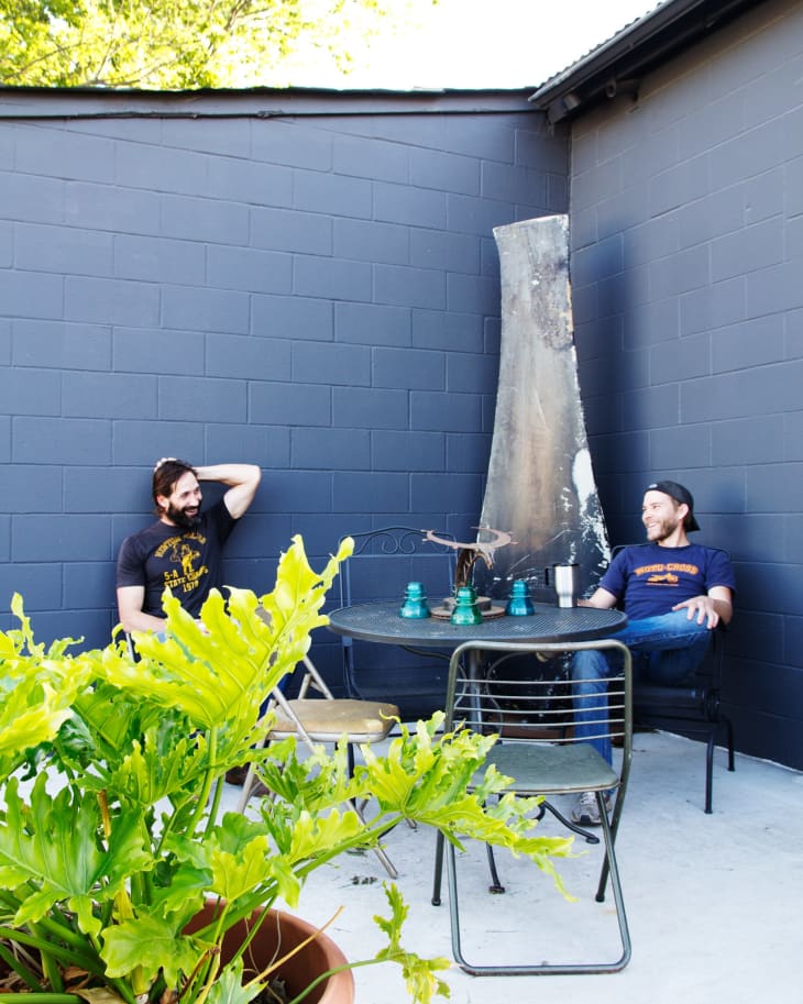 Two men sitting at a round metal table in a patio with a potted plant and dark blue walls.