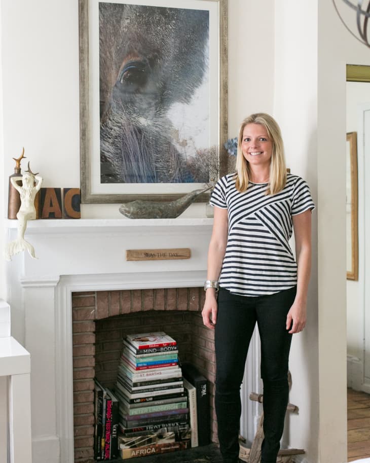 Woman in striped shirt standing by a fireplace with stacked books and large framed animal artwork above.