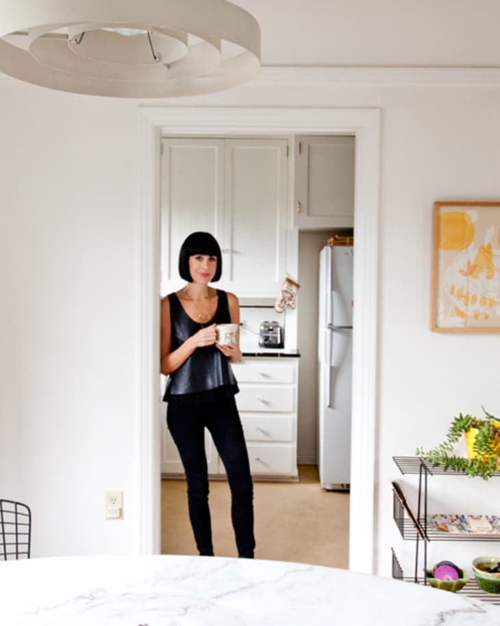 Woman in black top holding a mug, standing in a white kitchen doorway with a modern dining area.