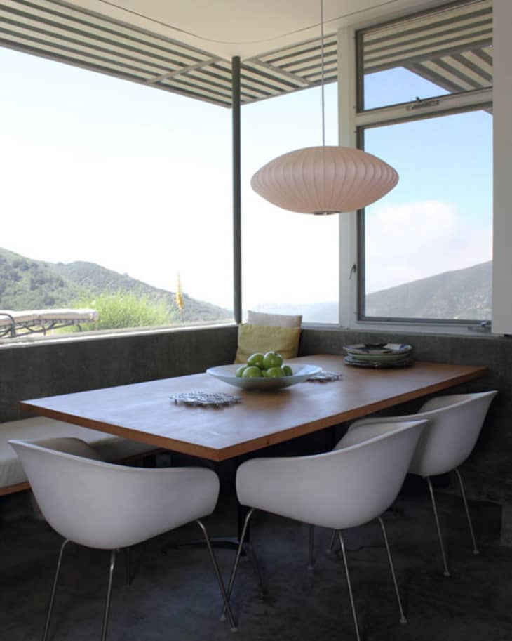 Dining area with a wooden table, white chairs, pendant light, and a bowl of green apples, overlooking a mountain view.