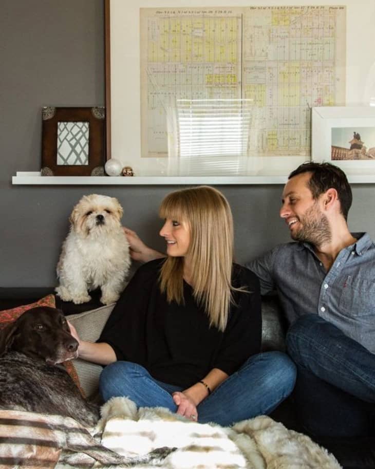 Couple sitting on a sofa with two dogs, framed map on wall shelf behind them.