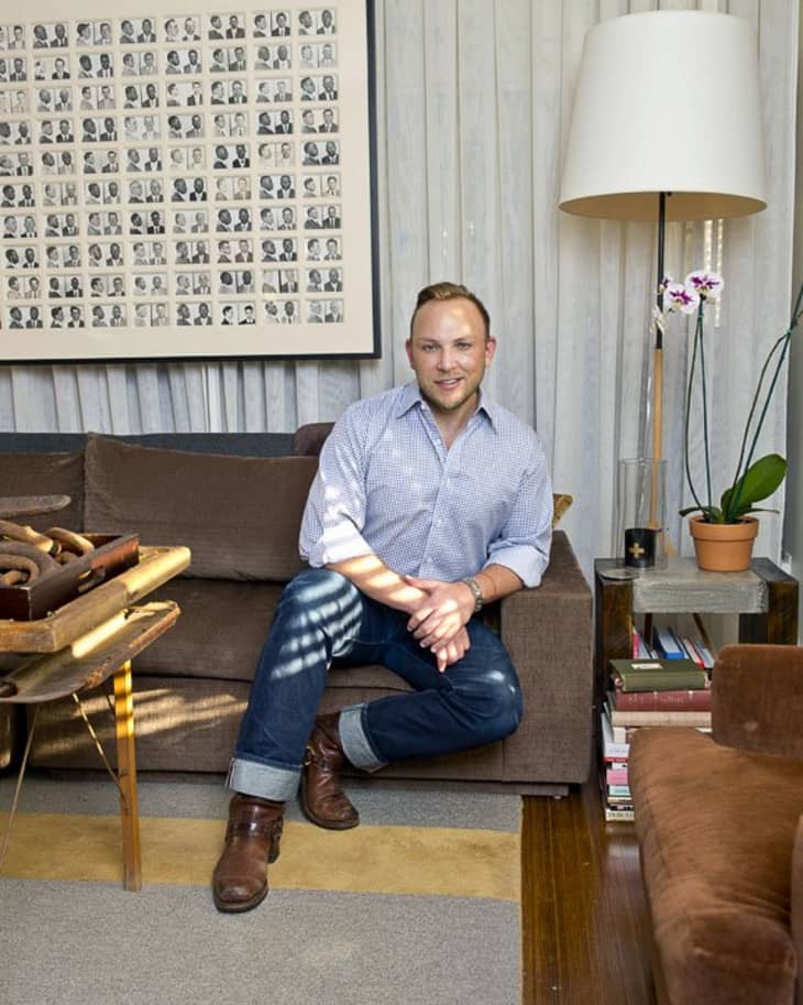 Man sitting on a brown sofa in a living room with a floor lamp, framed photo collage, and orchid plant.