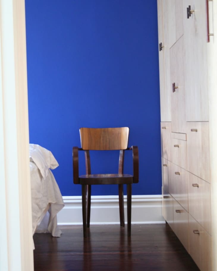 Wooden chair against a vibrant blue wall, beside a bed and light wood cabinets.