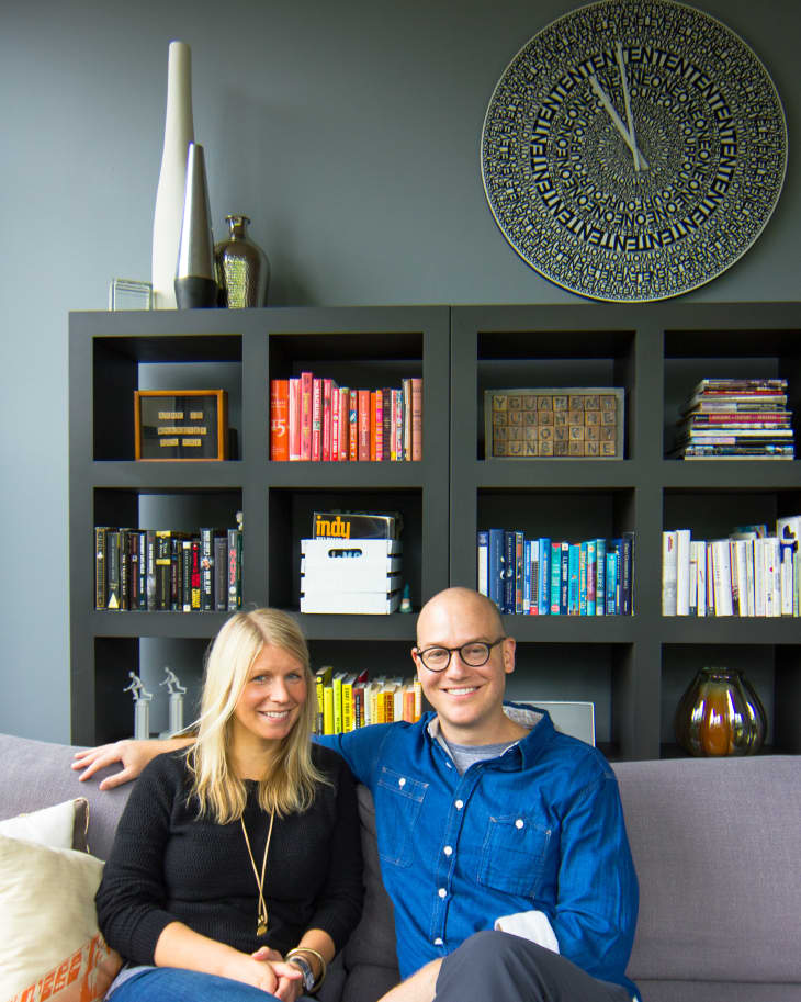 Couple sitting on a gray sofa in front of a bookshelf with colorful books and a large decorative clock.