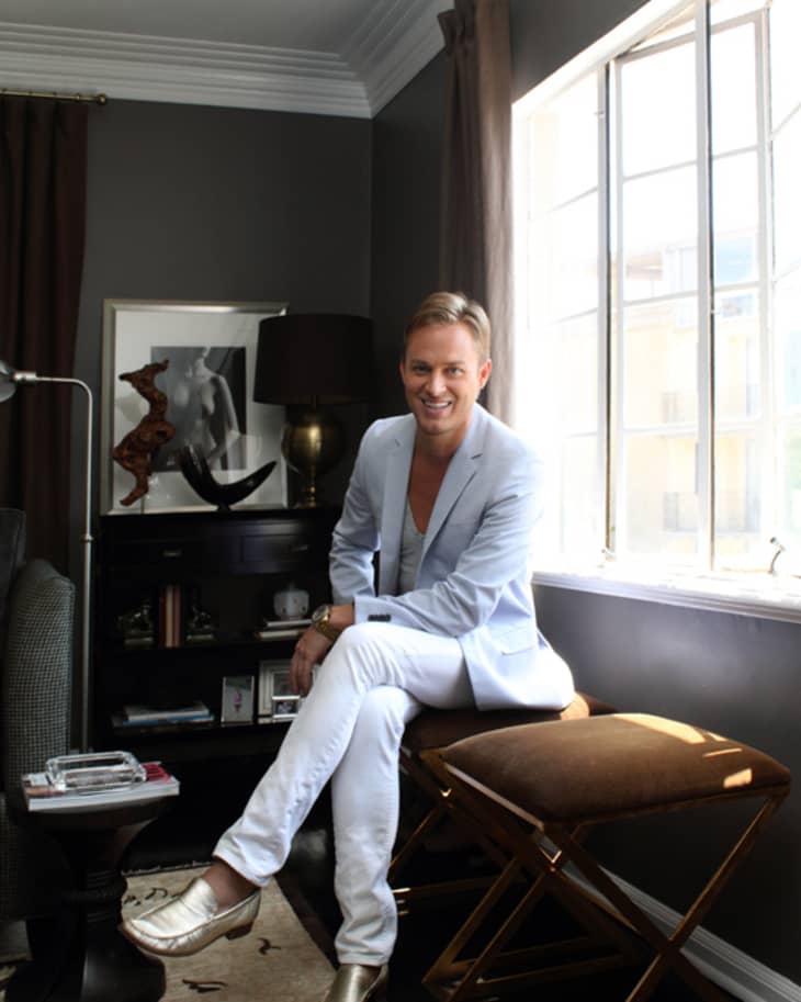 Man in light blue suit sitting on a stool by a window in a dark-toned room with art and decor.