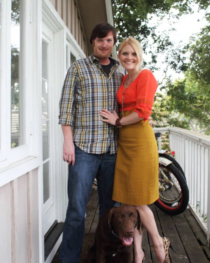 Couple standing on a wooden porch with a brown dog, next to a motorcycle, surrounded by trees.