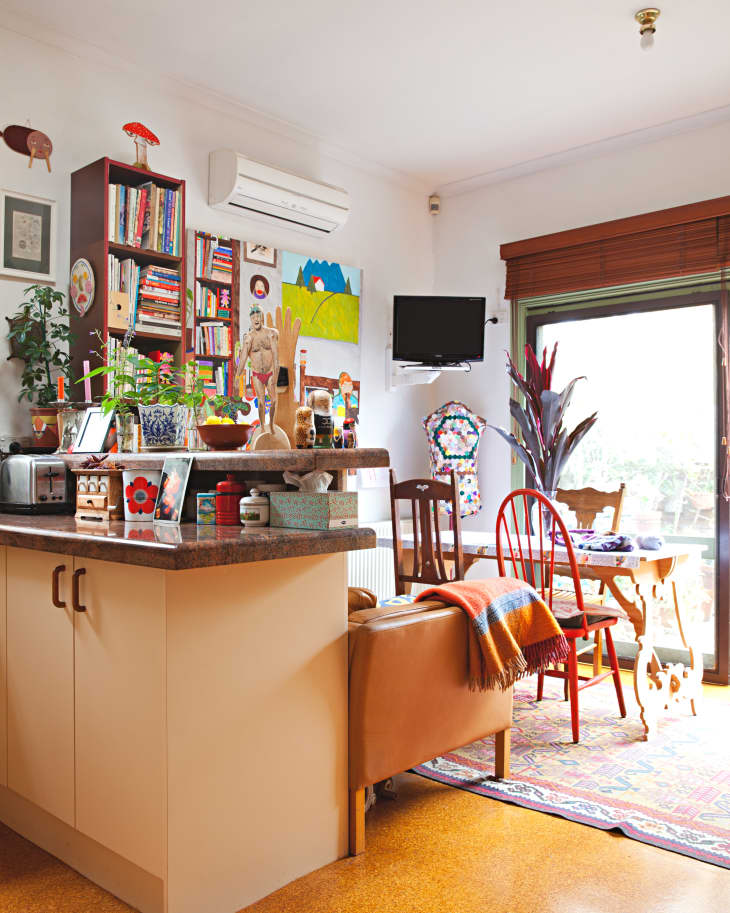 Cozy kitchen with bookshelves, colorful decor, wooden dining table, red chairs, and a small wall-mounted TV.