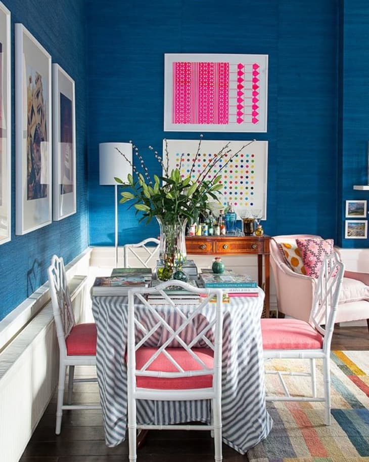 Dining room with blue walls, white chairs, pink cushions, striped tablecloth, and vibrant artwork.