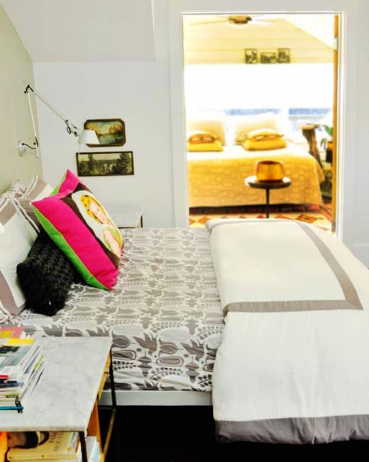 Bedroom with patterned bedding, colorful pillows, and a side table with books, leading to a sunlit room with a bed.