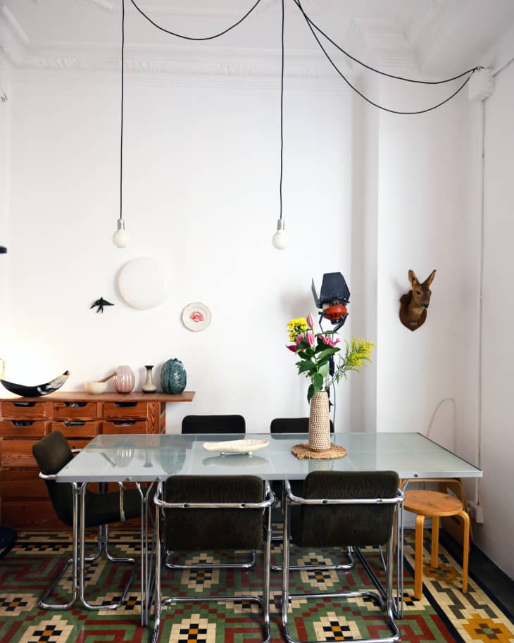 Dining room with glass table, six chairs, colorful geometric rug, pendant lights, and decorative vases on a wooden sideboard.