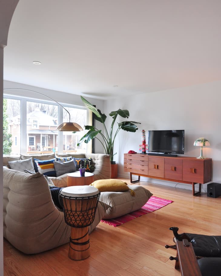Living room with a beige sectional sofa, colorful cushions, drum table, wooden TV stand, and large plant by the window.