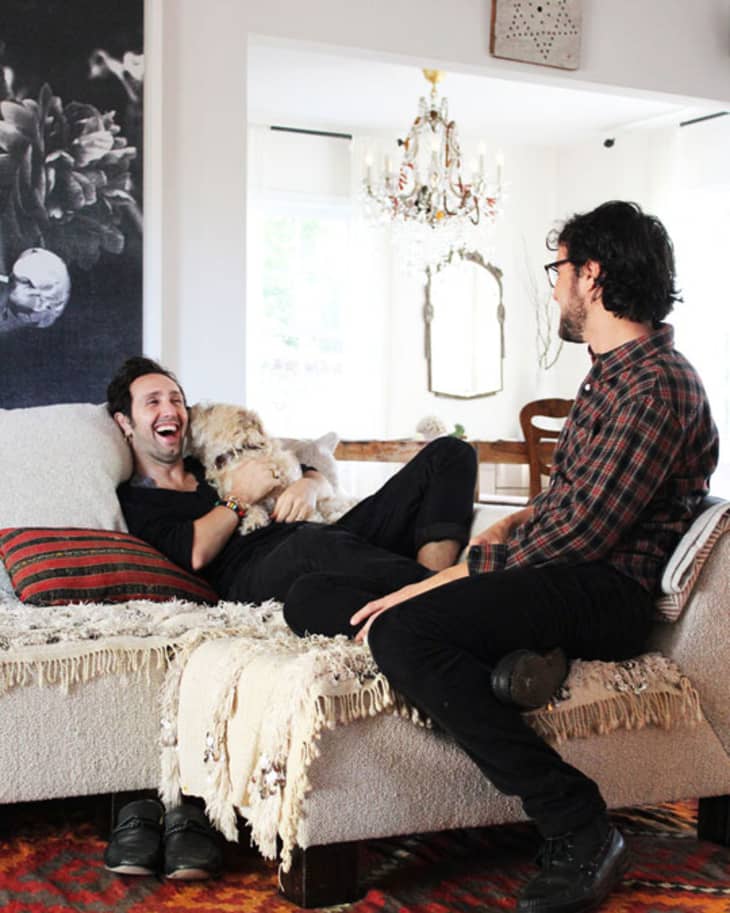 Two people laughing on a sofa with a dog, surrounded by cozy decor and a chandelier in a bright living room.