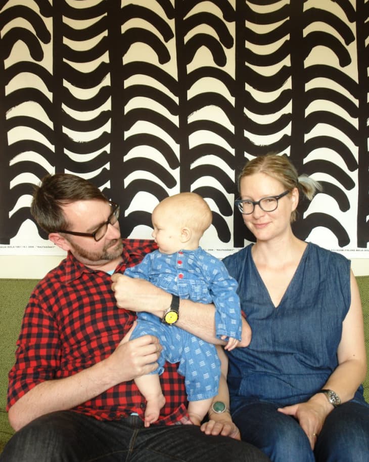 Man and woman sitting on a green sofa with a baby in blue overalls, against a bold black and white patterned wall.