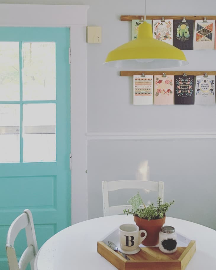 Dining area with a turquoise door, yellow pendant light, wall art, and a white table with a plant and mug.