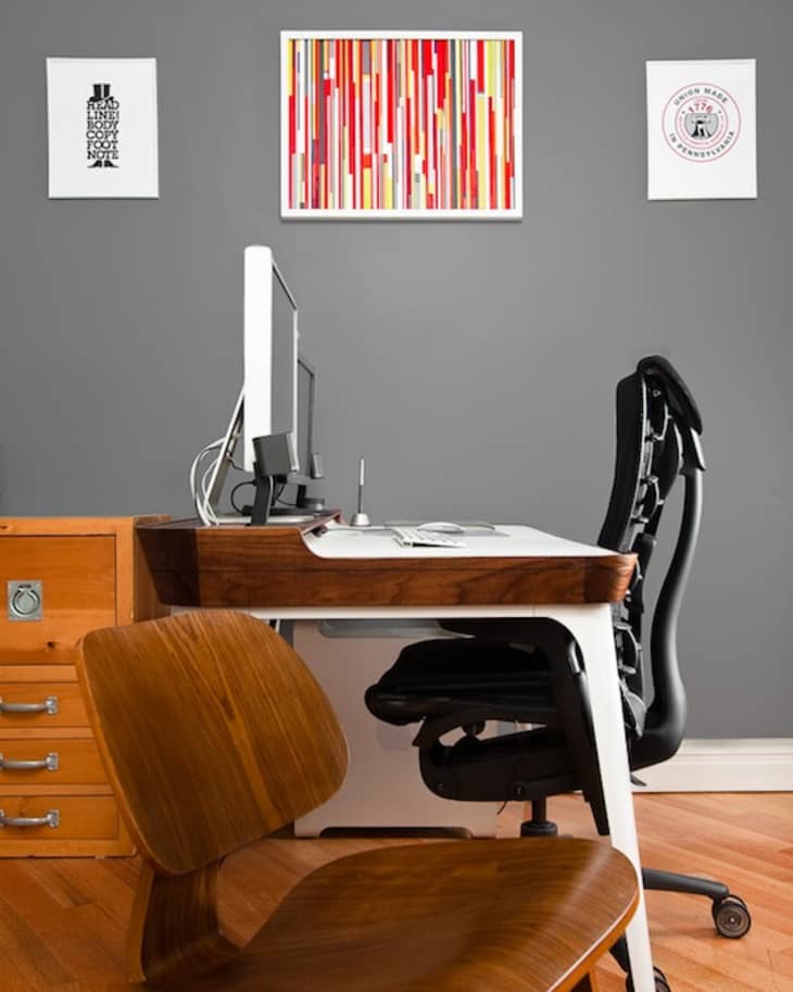 Home office with wooden desk, ergonomic chair, computer, and abstract art on gray wall.