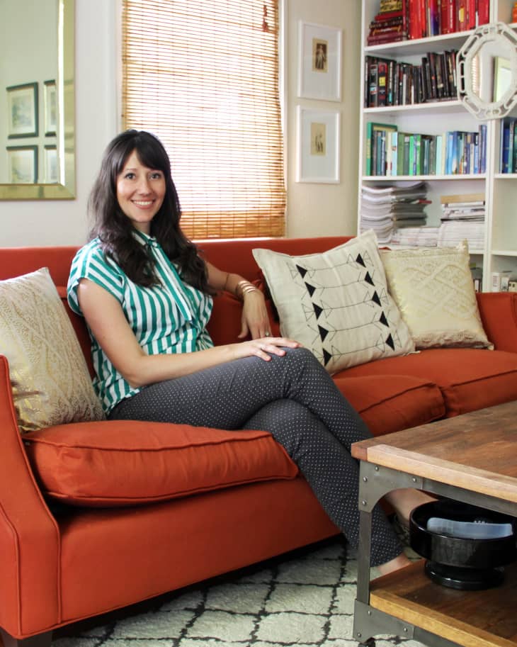 Woman sitting on an orange sofa with patterned cushions, next to a wooden coffee table and a bookshelf filled with colorful books.