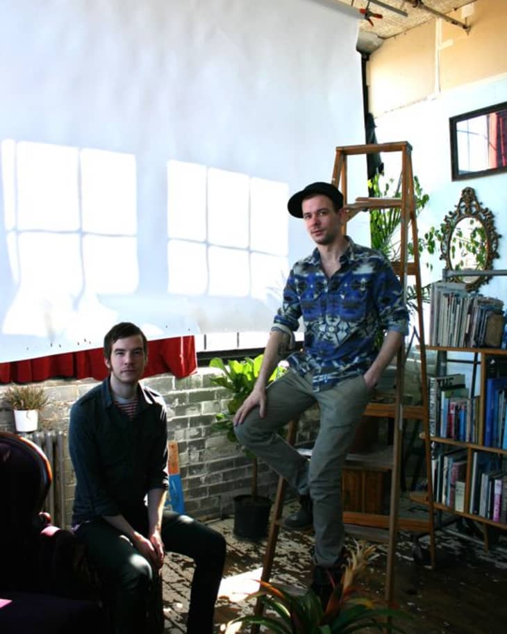 Two men in a sunlit room with a ladder, bookshelves, and plants.