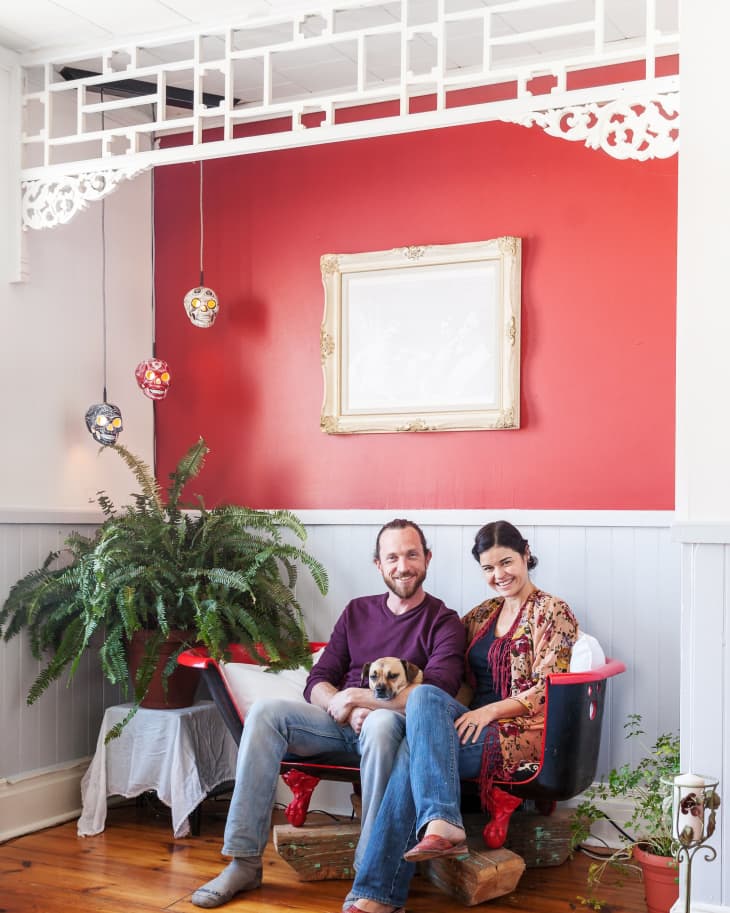 Couple sitting in a red clawfoot tub with a small dog, surrounded by plants, against a red accent wall with hanging lights.