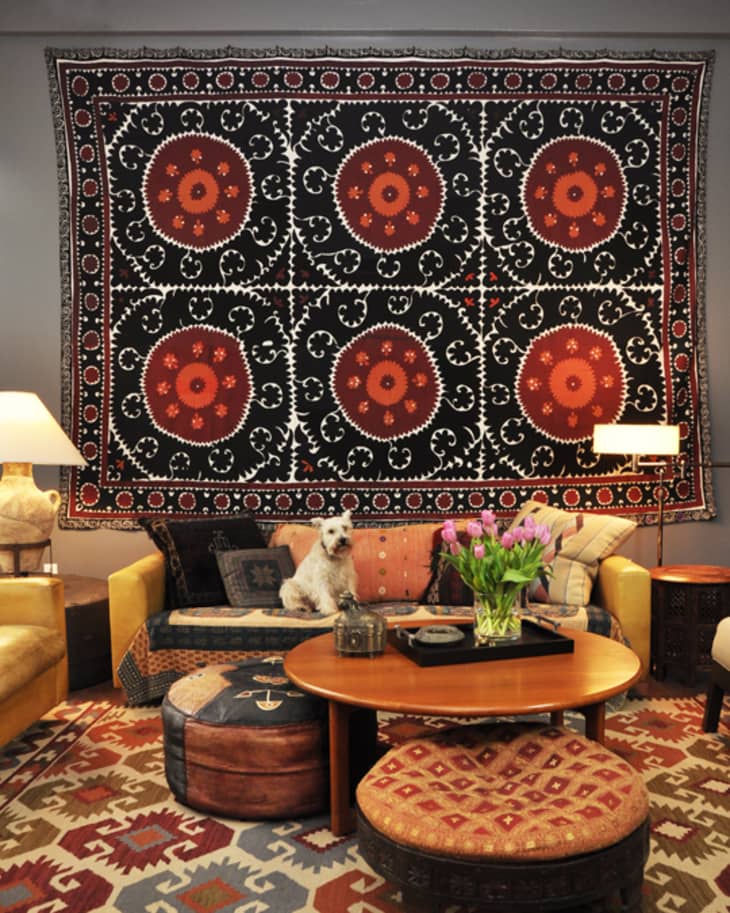 Living room with a large patterned tapestry, colorful rug, round wooden table, and a dog on the sofa.