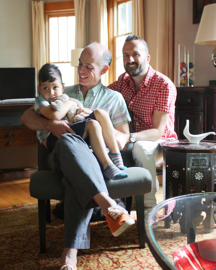 Two men and a child sitting on a chair in a cozy living room with a patterned rug and a glass coffee table.