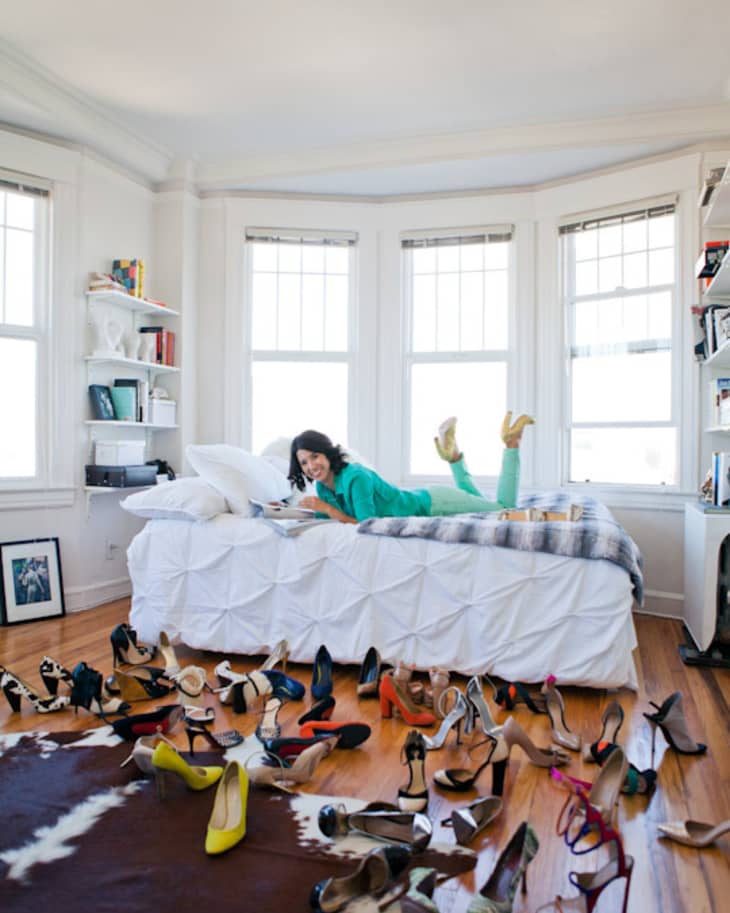 Woman in teal outfit on bed surrounded by various high heels in a bright room with large windows.