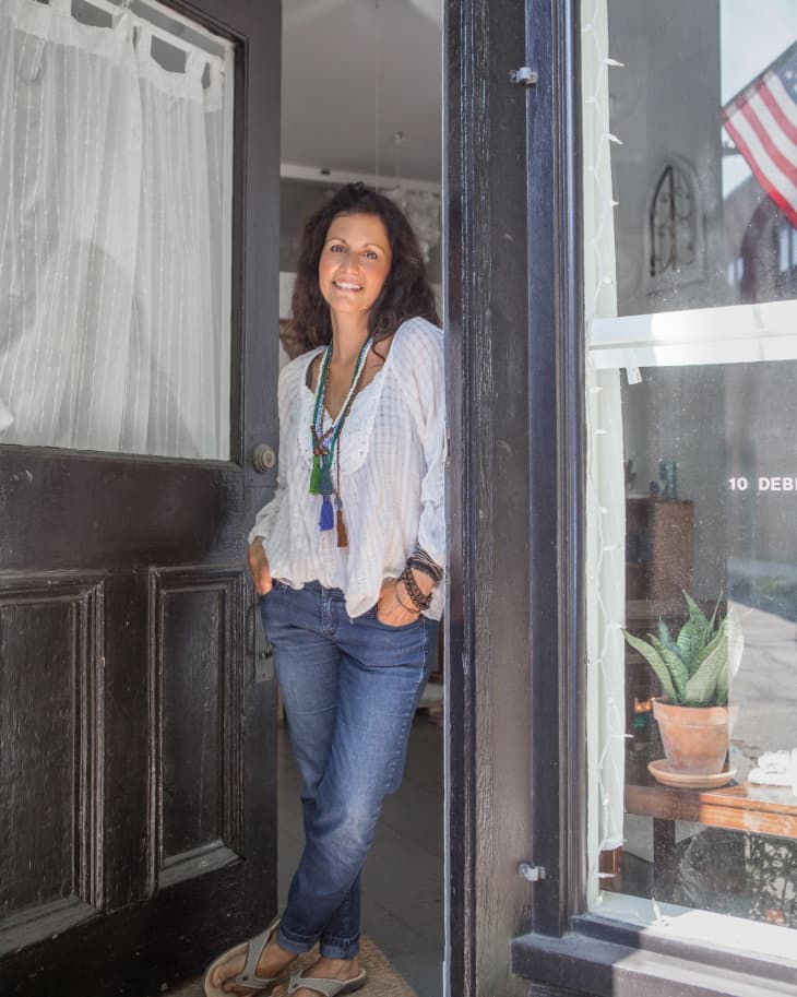 Woman in casual attire standing in doorway, smiling, with a potted plant visible through the window.