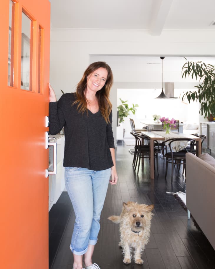 Woman in black top and jeans stands by an orange door with a small dog, leading to a dining area with plants.