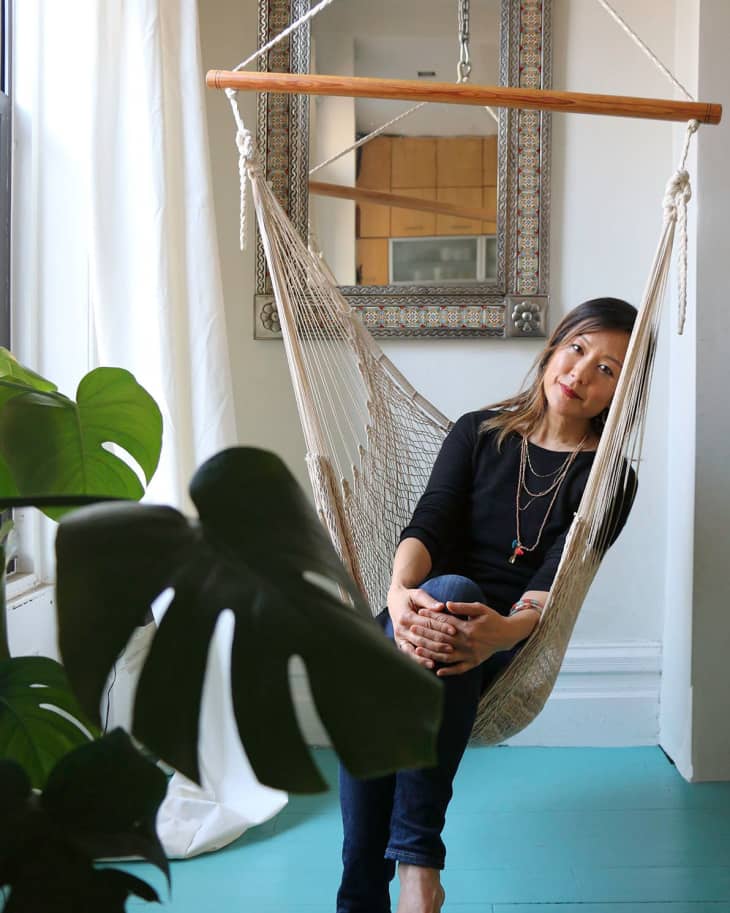Woman sitting in a hanging hammock chair, with a large mirror and plant in the background.