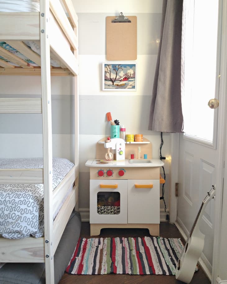 Bunk bed next to a play kitchen with colorful rug, clipboard, and wall art in a cozy room.