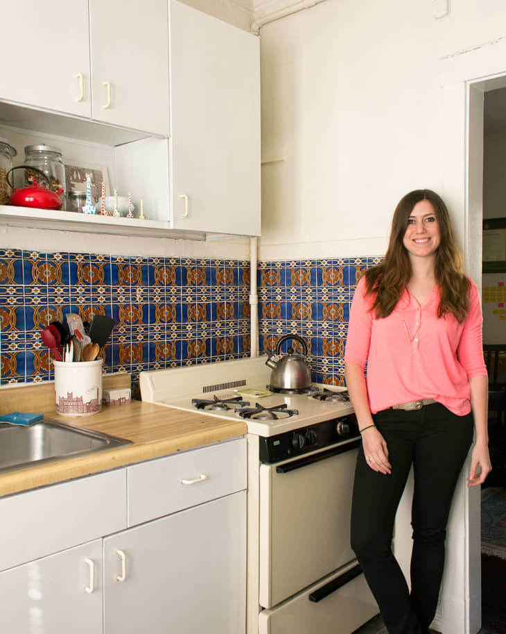 Woman in a kitchen with colorful tiled backsplash, standing by a stove with a kettle, wearing a pink top and black pants.