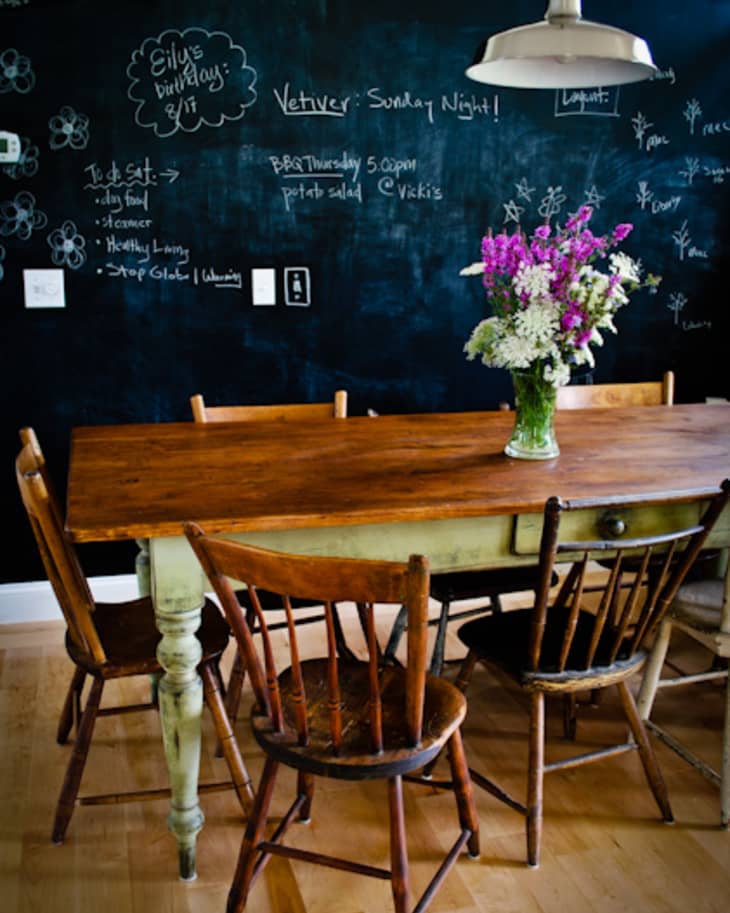 Rustic dining table with wooden chairs, chalkboard wall, and a vase of colorful flowers under a pendant light.