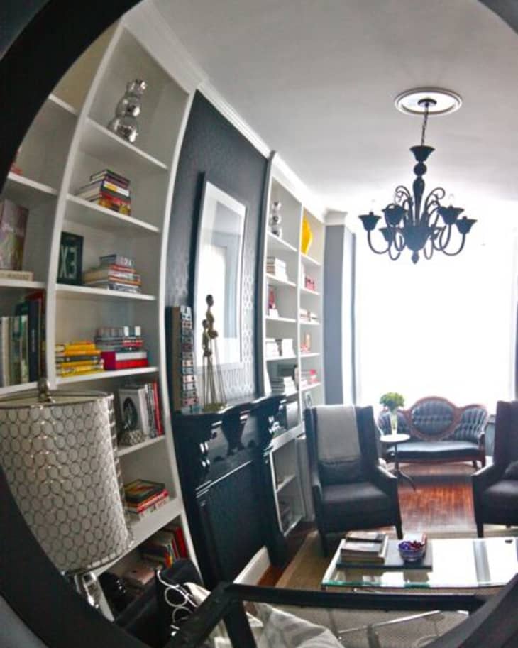 Modern living room with curved bookshelves, black chandelier, and armchairs reflected in a circular mirror.