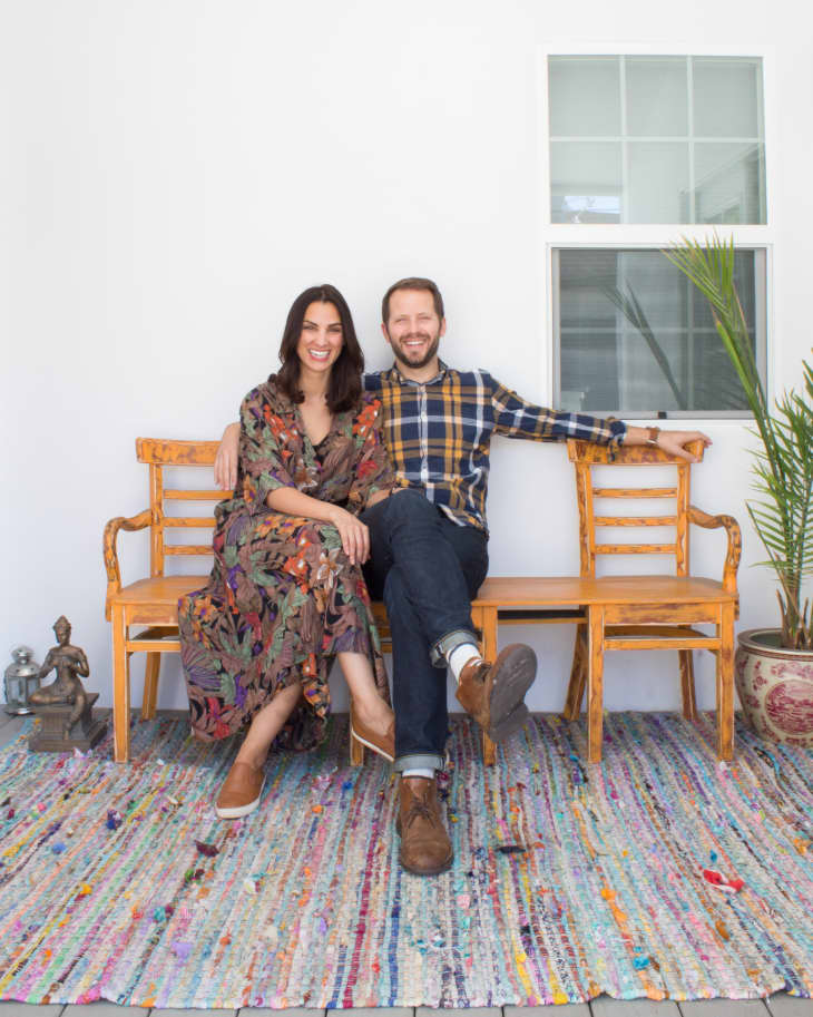 Couple sitting on a wooden bench with a colorful rug, potted plant, and decorative statue nearby.