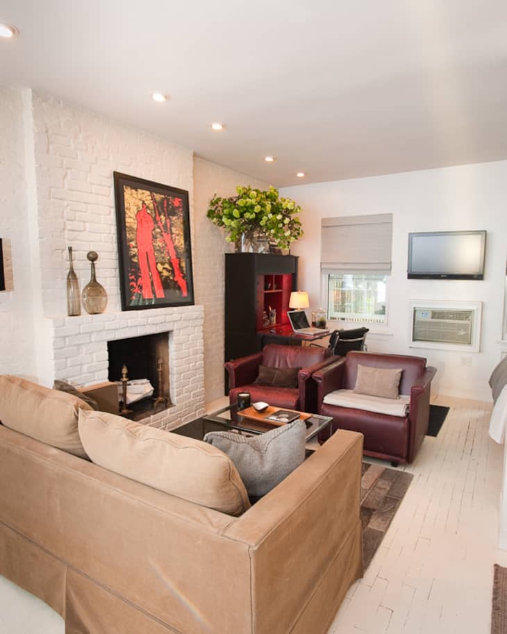 Living room with beige sofa, red armchairs, white brick fireplace, vibrant artwork, and a TV above an air conditioner.