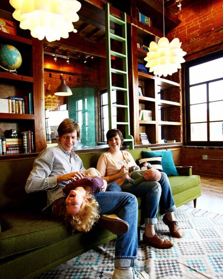 Family sitting on a green sofa in a cozy room with bookshelves, pendant lights, and a patterned rug.