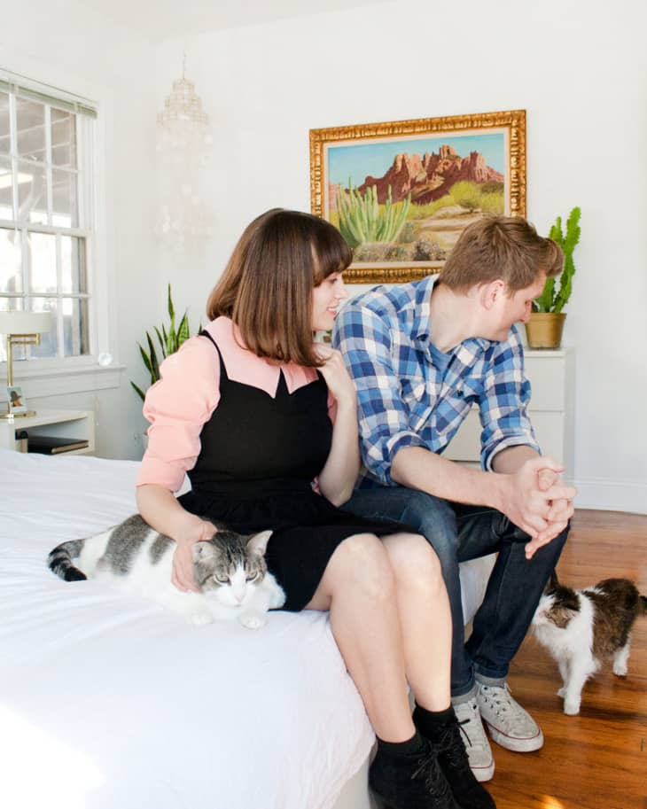 Couple sitting on a bed with two cats, surrounded by plants and a landscape painting in a bright room.