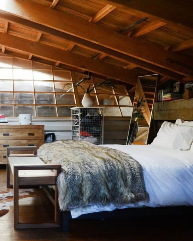 Cozy attic bedroom with sloped wooden ceiling, white bedding, fur throw, dresser, and a large mirror.