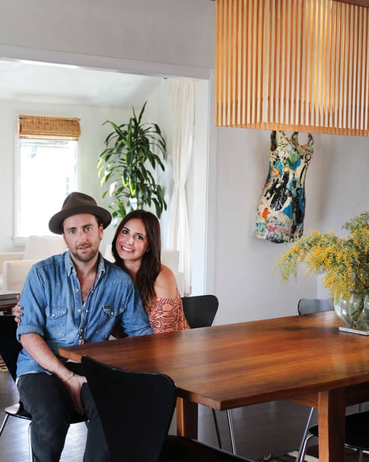 Couple sitting at a wooden dining table with black chairs, large plant, and a modern pendant light above.