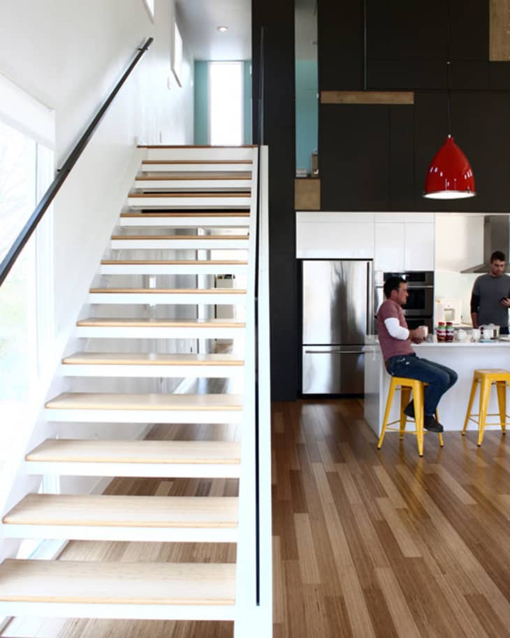 Modern kitchen with wooden floors, open staircase, two men at a counter with yellow stools, and a red pendant light.