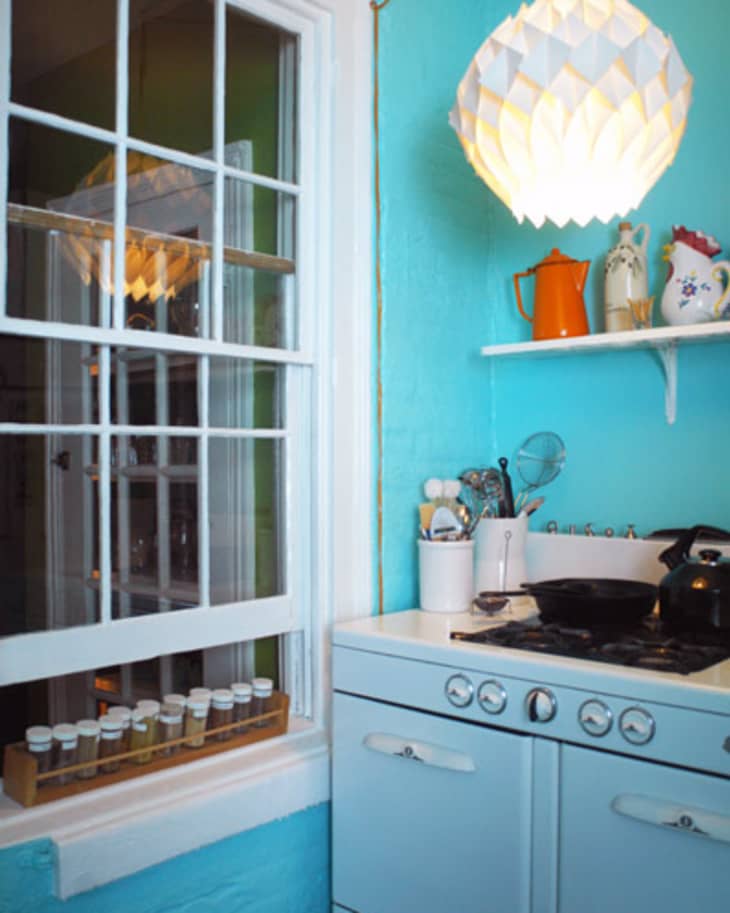 Retro kitchen with blue cabinets, white stove, hanging light, spice rack by window, and colorful teapots on a shelf.