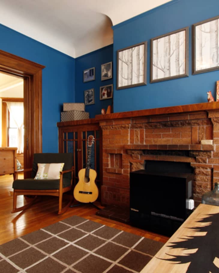 Living room with blue walls, brick fireplace, guitar, framed art, and a wooden chair with a cushion.