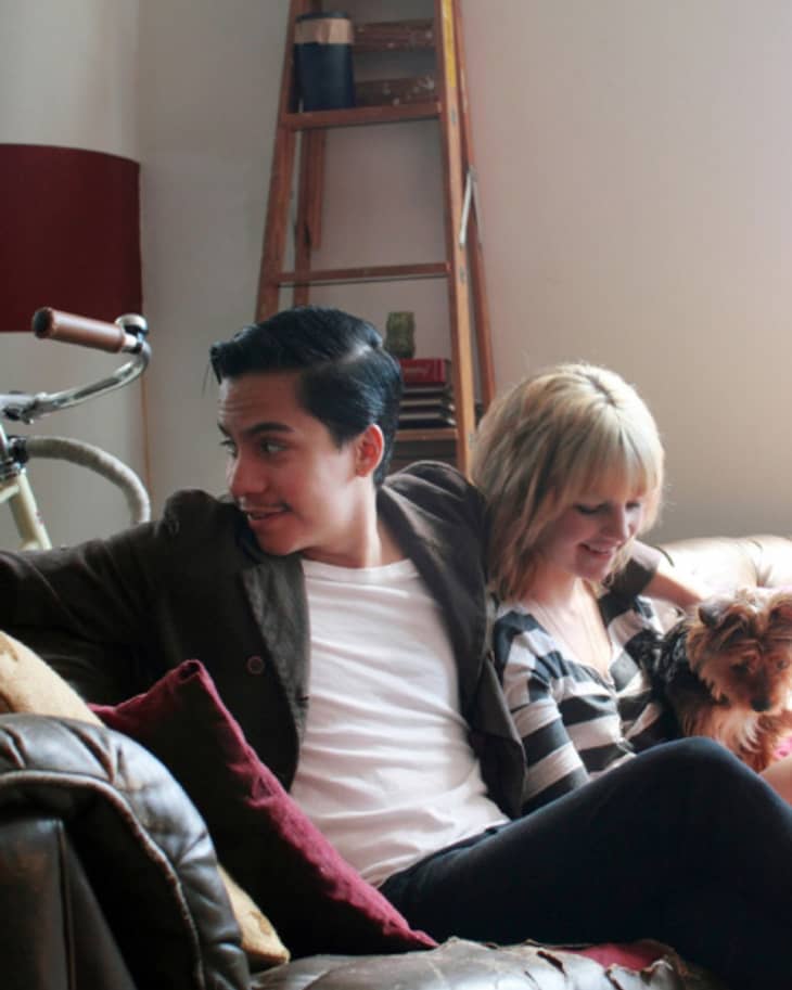 Couple sitting on a leather sofa with a small dog, bicycle, and ladder in the background.
