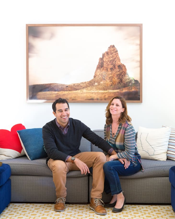 Couple sitting on a gray sofa with colorful cushions, beneath a large framed mountain landscape photo.