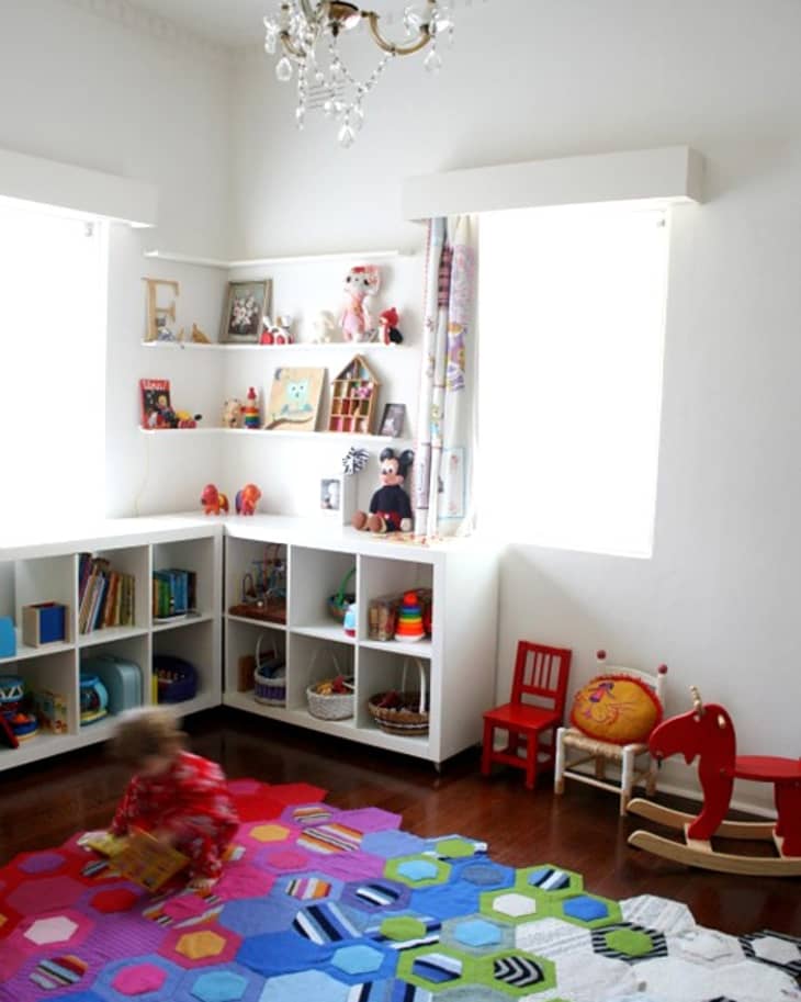 Child playing on colorful hexagon-patterned rug in a bright playroom with shelves of toys and books.