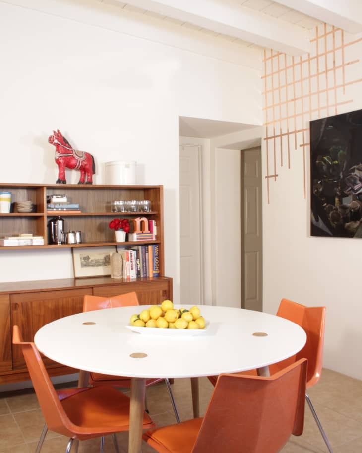Dining room with white round table, orange chairs, wooden cabinet, red elephant figurine, and bowl of lemons.